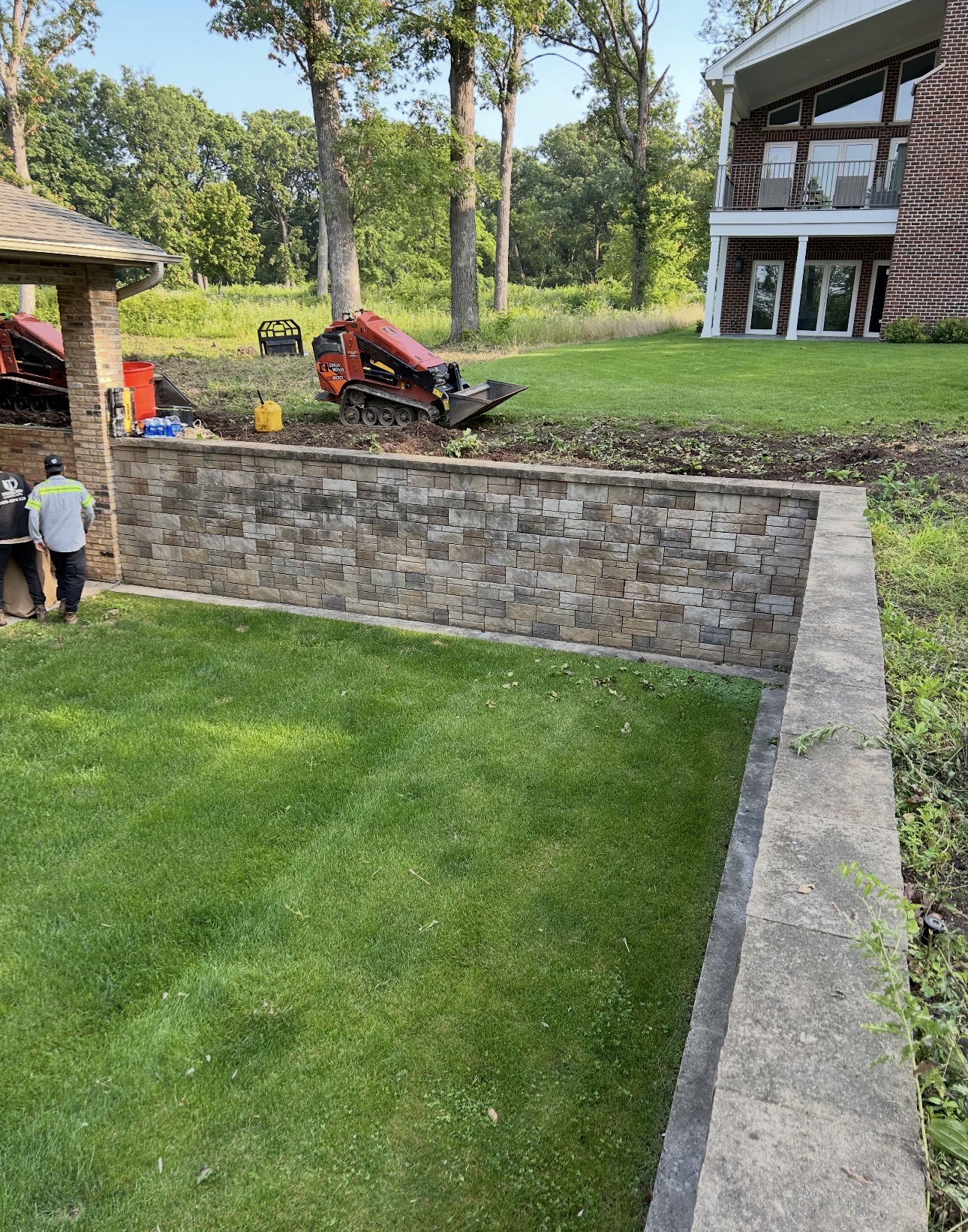 Long multi-tone block retaining wall with capstones separating a manicured lawn from a wooded slope