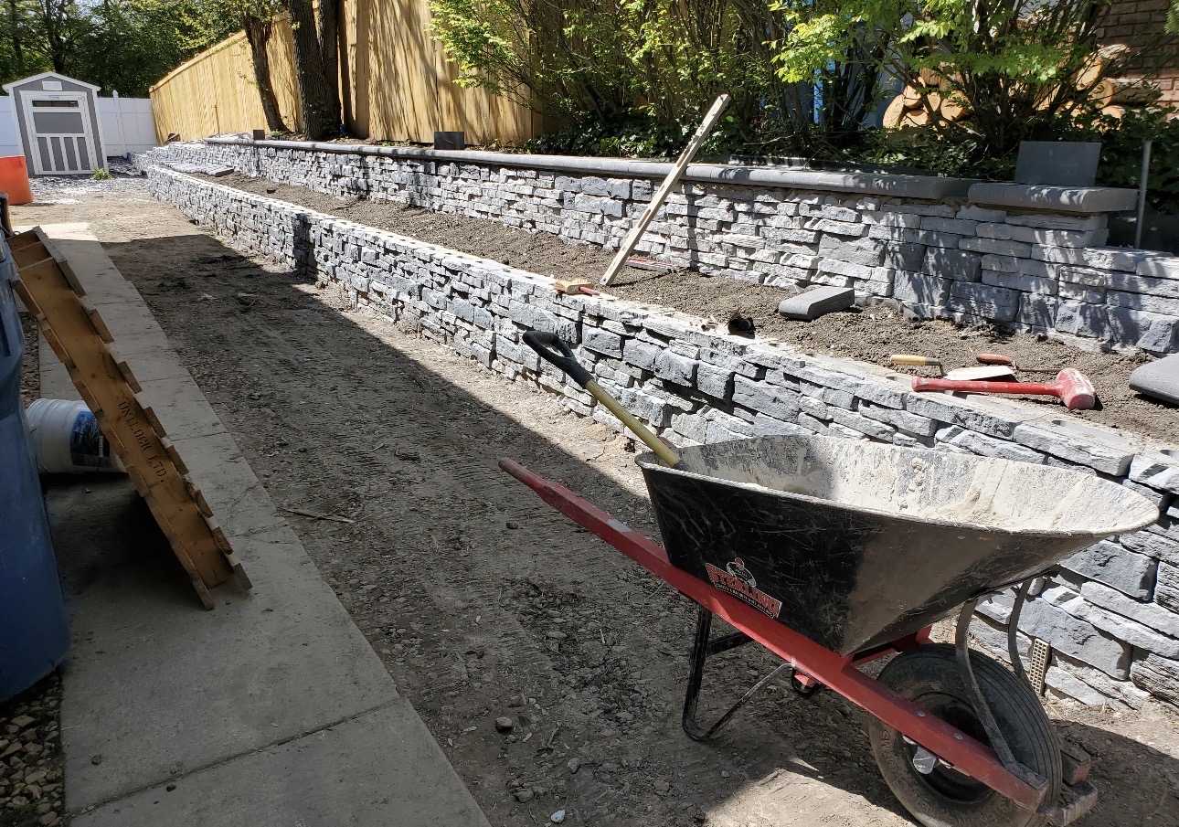 Long stacked stone wall mid-build with wheelbarrow and tools on the job site