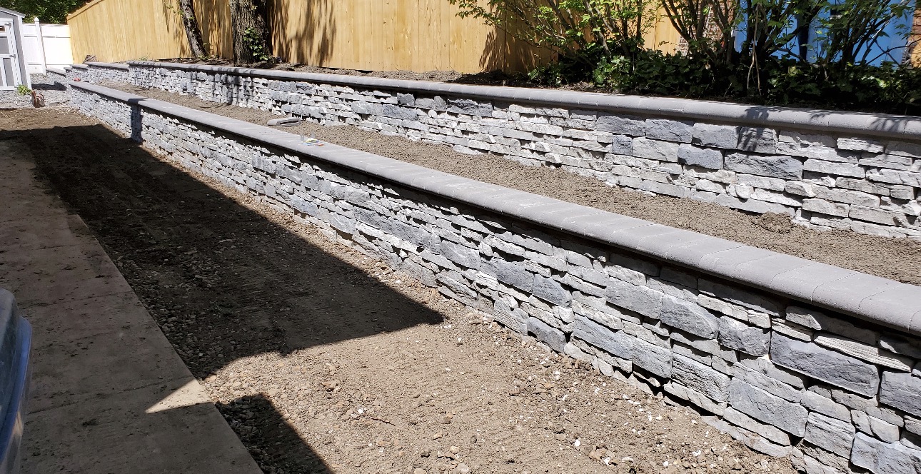 Two-tier grey stacked stone retaining wall with capstones along a backyard fence line